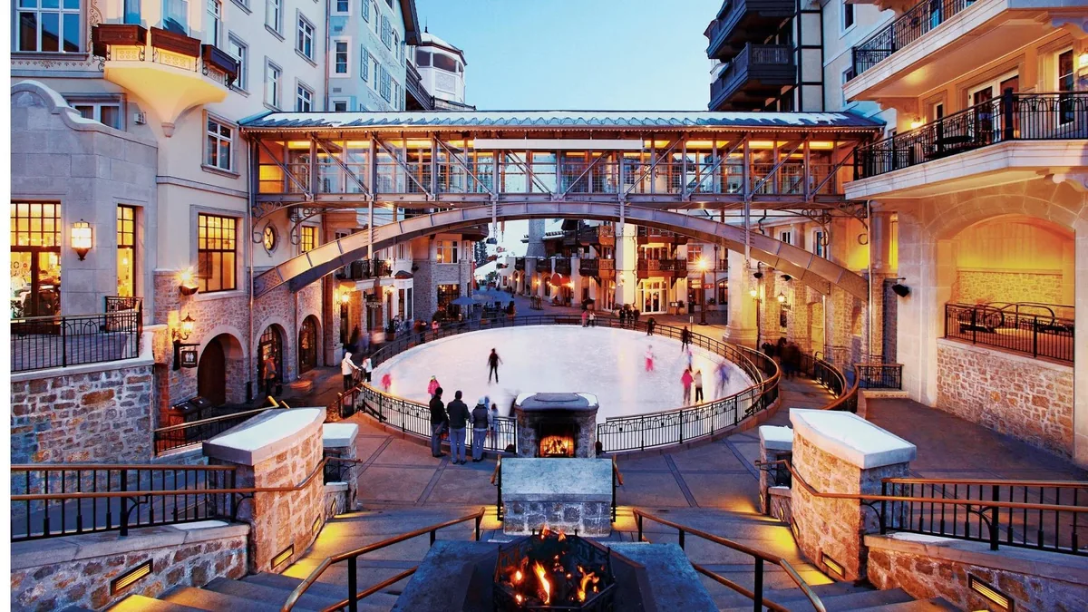 Skaters on the open-air ice rink in the plaza at dusk, with a covered walkway overhead and a fire pit in the foreground