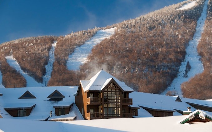 Snow-covered lodge roofs in front of groomed ski trails cut through a forested mountainside