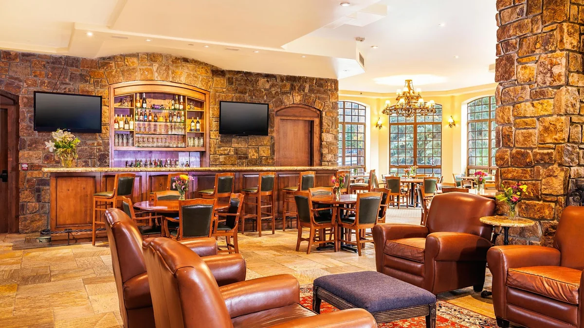 Lounge with a stone-faced bar, two wall-mounted televisions, wooden tables and leather armchairs in front of tall arched windows