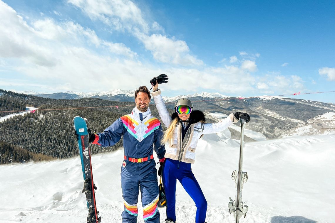 Two skiers standing on a snowy ridge with skis planted upright, forested hills and a distant snowy mountain range behind them